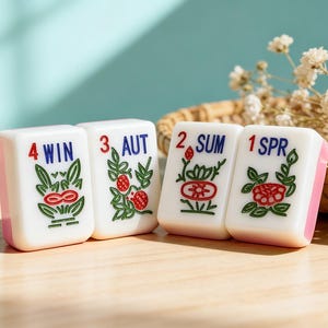 Peut inclure: Quatre tuiles de Mahjong blanches avec des bords roses. Chaque tuile affiche une saison : WIN, AUT, SUM et SPR, avec des illustrations rouges, vertes et bleues. Un panier en osier et des fleurs sont en arri&egrave;re-plan, sur une surface en bois.