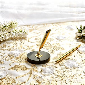 May include: A gold pen and pen holder set. The pen holder has a black base and a gold pen rest. The pen is gold and sleek. The set is on a white and gold floral patterned surface.