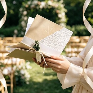 May include: A tan cardstock booklet with the word Vows printed in white script, featuring a textured white paper overlay with a torn edge, tied with a white string, a small green sprig, and a rectangular tag with the word Thanks printed on it.