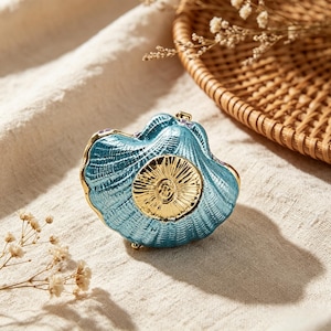 May include: A decorative, blue seashell-shaped trinket box with gold trim and a gold center design. The box is resting on a cream-colored fabric, with a wicker basket and dried flowers in the background.