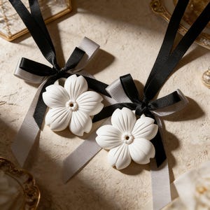 Puede incluir: Dos adornos blancos con forma de flor con cintas negras y grises. Las flores tienen diseños detallados de pétalos y un pequeño agujero en el centro. Las cintas están atadas en lazos, creando una apariencia delicada y elegante. Estos adornos podrían usarse para decoración o como accesorios.