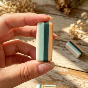 May include: A rectangular, smooth, multi-colored block featuring vertical stripes of light mint green, dark forest green, and soft peach. The object has a glossy finish and rounded edges, with a flat, uniform shape that is held between fingers.