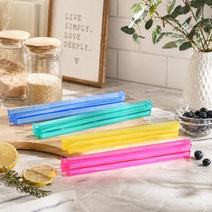 May include: Four colorful plastic bag sealers in blue, green, yellow, and pink are arranged on a wooden cutting board. The sealers are next to a bowl of blueberries, lemon slices, and a framed sign that reads "LIVE SIMPLY, LOVE DEEPLY."