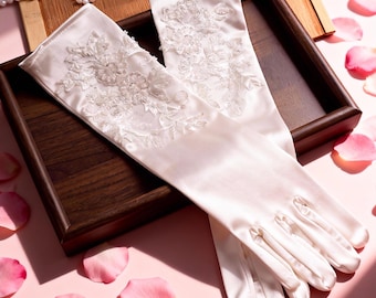 May include: Ivory satin gloves with floral lace embellishments, presented on a wooden tray. Pink rose petals and a pearl necklace surround the gloves. The image includes the text "My Heart Never Stops Beating for You" at the bottom.