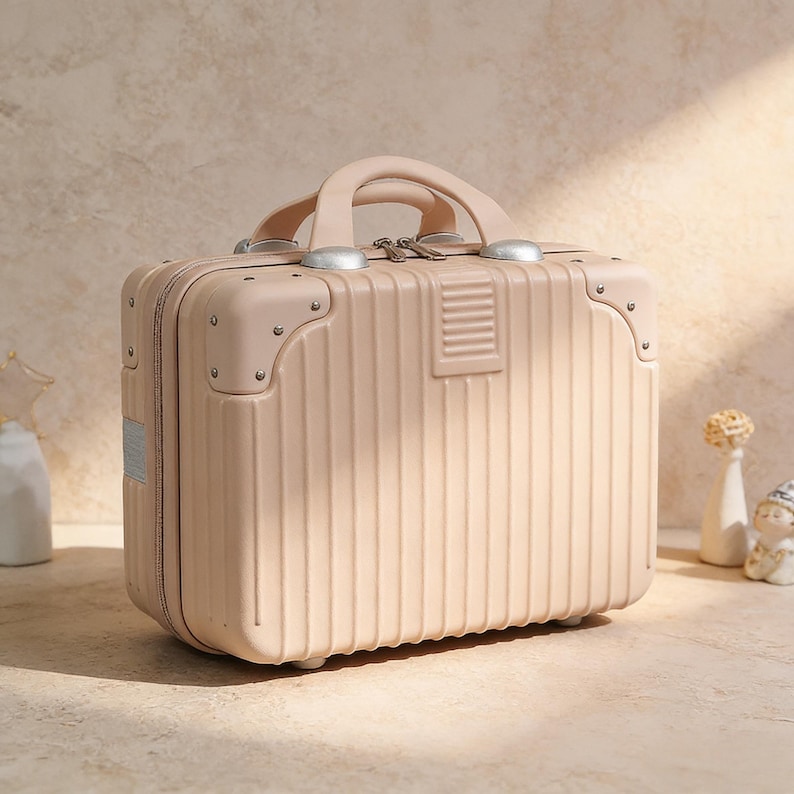 May include: A blush-colored, small suitcase with a handle and silver-toned hardware. The suitcase has vertical ribbed detailing and rounded corners. The handle is arched, and the suitcase has a rectangular shape. The suitcase is sitting on a light-colored surface.