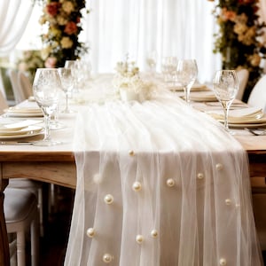 May include: A beautifully set dining table with white plates, silverware, and crystal wine glasses. A sheer white table runner adorned with faux pearls drapes over the table. Floral arrangements and white chairs complete the elegant setting.