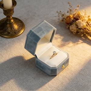 May include: A light blue velvet ring box with a gold clasp, open to reveal a diamond ring. The ring box is octagonal and lined with white fabric. A gold candle holder and dried flowers are in the background.