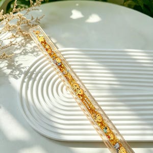 Könnte beinhalten: Ein durchsichtiges Kunststofflineal mit einem goldenen Glitzerdesign und Cartoonfiguren. Das Lineal hat schwarze Messmarkierungen und ist mit kleinen, lächelnden, bärenartigen Figuren verziert. Das Lineal ist über eine weiße, geriffelte Oberfläche geneigt.