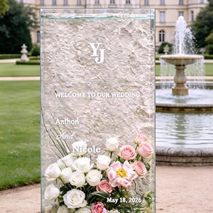 May include: A clear glass wedding sign with white text that reads "Welcome to our Wedding" and the names "Anthon and Nicole". The sign is decorated with white and pink roses and the date "May 18, 2026". The sign is in front of a fountain.