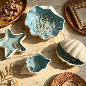 May include: A collection of ceramic seashell-shaped trinket dishes in shades of blue and white, filled with jewelry. A starfish-shaped dish and a conch shell dish are also present, all arranged on a wooden surface.