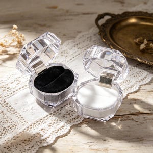 May include: Two clear, faceted, hinged jewelry boxes. One box has a black velvet interior with two slots, the other has a white interior. The boxes are open, revealing their contents. The boxes are on a white lace surface.