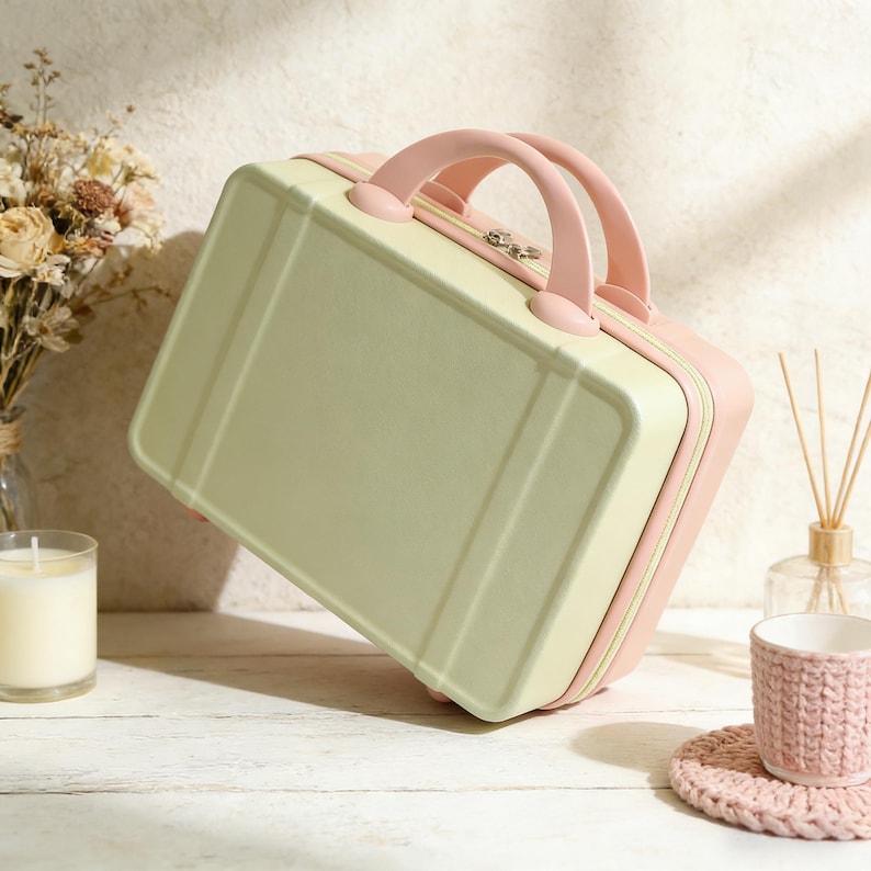 May include: A small, rectangular suitcase in pastel green and pink. The suitcase has a rounded top with a pink handle and trim. The handle is arched and the suitcase has a silver clasp. The suitcase is sitting on a white surface.