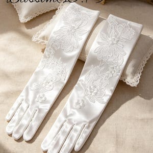 May include: Two white satin gloves with intricate floral lace detailing. The gloves are long, reaching up the arm, and are presented on a neutral background. The image includes the text "Blossom (15.7)".