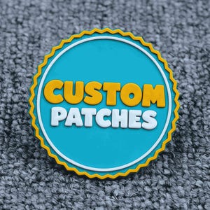 Könnte beinhalten: Ein runder Aufnäher mit türkisfarbenem Zentrum und den Worten "CUSTOM PATCHES" in Weiß und Gelb. Der Aufnäher hat einen weißen Rand und einen gelben, gezackten Rand. Der Aufnäher liegt auf einer grauen, strukturierten Oberfläche.