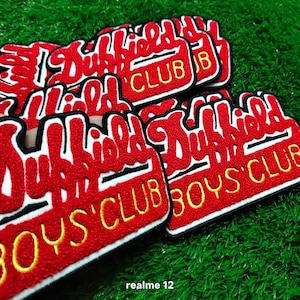 May include: Several red and white embroidered patches with the text "Dubfield Boys Club". The patches have a black border and are displayed on a green surface, with a textured appearance.