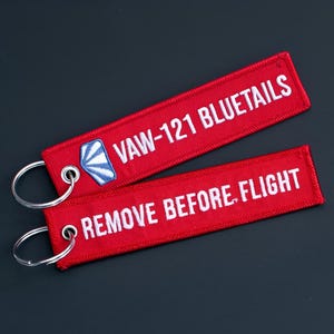 Puede incluir: Dos llaveros de tela roja con texto blanco. Uno dice "VAW-121 BLUETAILS" con un emblema azul y blanco. El otro dice "REMOVE BEFORE FLIGHT". Ambos tienen llaveros plateados.