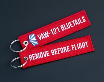 Könnte beinhalten: Zwei rechteckige Schlüsselanhänger aus rotem Stoff mit silbernen Metallringen. Einer zeigt den weißen Sticktext VAW-121 BLUETAILS und ein blau-weißes Emblem. Der andere zeigt den weißen Sticktext REMOVE BEFORE FLIGHT.
