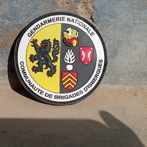 K&ouml;nnte beinhalten: Runder Aufn&auml;her mit den Worten "GENDARMERIE NATIONALE" und "COMMUNAUTE DE BRIGADES D'ISBERGUES". Der Aufn&auml;her zeigt einen schwarzen L&ouml;wen, ein Schild und andere Embleme in Gelb, Rot und Schwarz.
