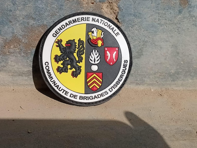 K&ouml;nnte beinhalten: Runder Aufn&auml;her mit den Worten "GENDARMERIE NATIONALE" und "COMMUNAUTE DE BRIGADES D'ISBERGUES". Der Aufn&auml;her zeigt einen schwarzen L&ouml;wen, ein Schild und andere Embleme in Gelb, Rot und Schwarz.