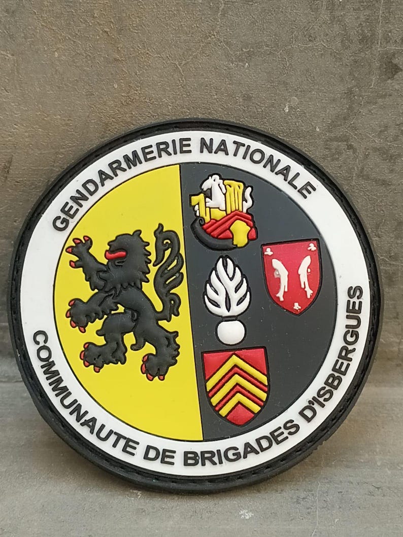 K&ouml;nnte beinhalten: Ein runder Aufn&auml;her mit den Worten "GENDARMERIE NATIONALE" und "COMMUNAUTE DE BRIGADES D'ISBERGUES". Der Aufn&auml;her zeigt einen schwarzen L&ouml;wen auf gelbem Grund und weitere Embleme in Rot, Wei&szlig; und Schwarz.