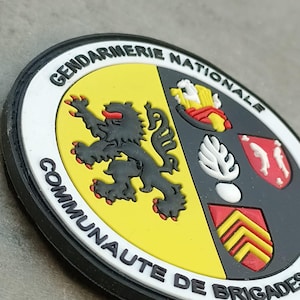 K&ouml;nnte beinhalten: Ein kreisf&ouml;rmiger Aufn&auml;her mit dem wei&szlig;en Text "GENDARMERIE NATIONALE COMMUNAUTE DE BRIGADES". Der Aufn&auml;her zeigt einen schwarzen L&ouml;wen auf gelbem Grund und ein Schild mit roten, wei&szlig;en und gelben Elementen. Der Rand ist schwarz.