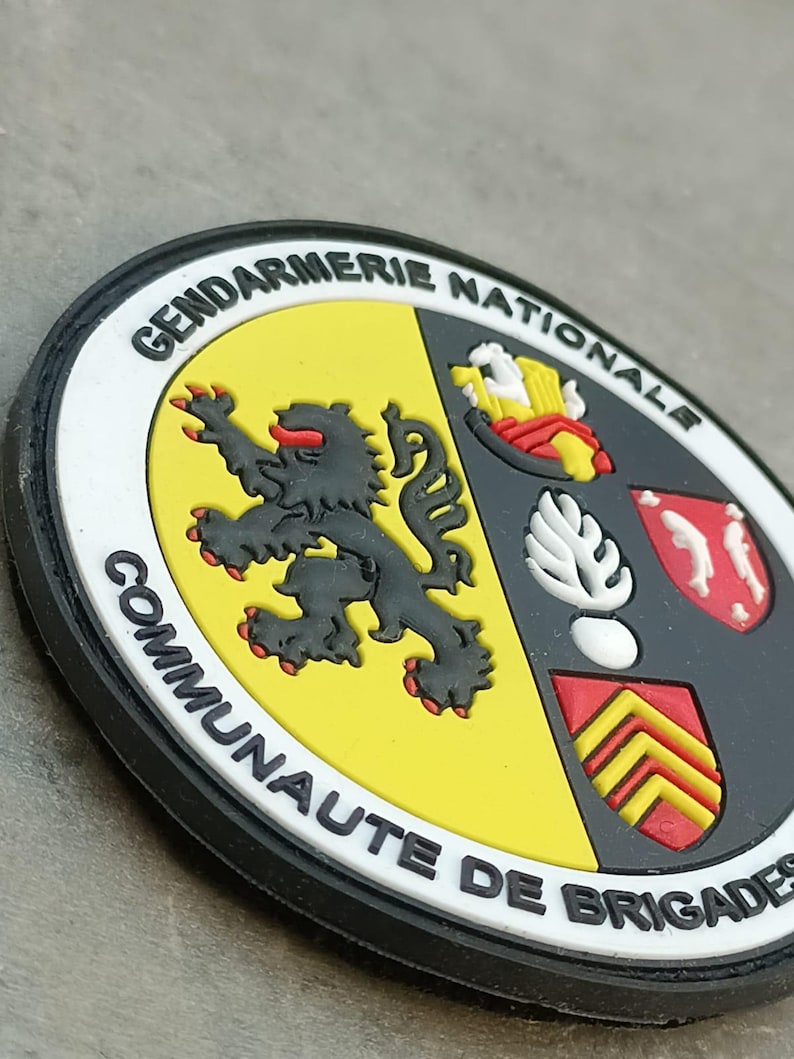 K&ouml;nnte beinhalten: Ein kreisf&ouml;rmiger Aufn&auml;her mit dem wei&szlig;en Text "GENDARMERIE NATIONALE COMMUNAUTE DE BRIGADES". Der Aufn&auml;her zeigt einen schwarzen L&ouml;wen auf gelbem Grund und ein Schild mit roten, wei&szlig;en und gelben Elementen. Der Rand ist schwarz.