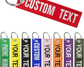 Könnte beinhalten: Eine Sammlung rechteckiger, bestickter Stoffschlüsselanhänger in verschiedenen Farben wie Rot, Grün, Gelb, Schwarz, Blau, Pink, Orange, Lila und Oliv. Jeder Schlüsselanhänger hat einen silbernen Metallring und eine weiße Stickerei.