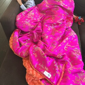 以下が含まれることがあります： 茶色のソファに掛けられた、金とターコイズの模様が入った鮮やかなピンク色の毛布。毛布の裏側はオレンジ色のぬいぐるみです。毛布は2人の子供を覆うのに十分な大きさです。