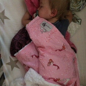 Puede incluir: Un niño dormido está acurrucado en una cuna, cubierto con una manta rosa adornada con ilustraciones de sirenas. La manta tiene un reverso blanco y esponjoso. También se ve una almohada de felpa rosa y un chupete.
