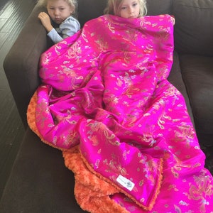 Könnte beinhalten: Eine leuchtend pinkfarbene Decke mit einem goldenen Muster aus Vögeln und Wolken, kombiniert mit einer weichen, orangefarbenen Plüschrückseite. Die Decke liegt auf einem braunen Sofa. Ein kleines Etikett ist sichtbar.