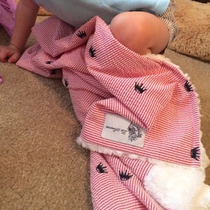May include: A red and white striped baby blanket with a white fluffy trim and black crown embroidery. The blanket has a white tag with the text "Little & Brave" on it.