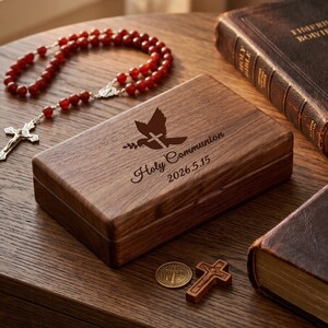 May include: A wooden box with a dove and cross design, the words "Holy Communion" and the date 2026.5.15 are engraved on the lid. A rosary with red beads, a small wooden cross, a coin, and a leather-bound book are also present.