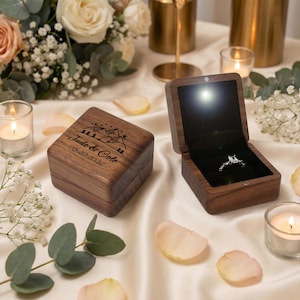 May include: Two wooden ring boxes, one closed and one open, sit on a white satin surface. The open box reveals a diamond ring. The closed box is engraved with a mountain scene and the names "Jude & Cole" and the date "05-20-2026".