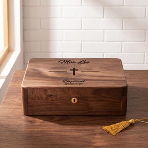 Könnte beinhalten: Eine rechteckige Holzkiste mit Schloss und goldenem Schlüsselloch. Die Kiste ist mit dem Namen "Mia Lee", einem Kreuz und dem Wort "Baptized" mit dem Datum "2026.5.15" graviert. Rechts ist eine goldene Quaste.