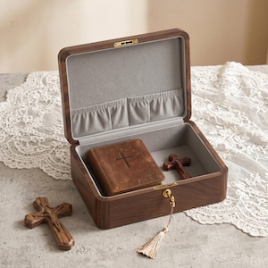 May include: A wooden keepsake box with a hinged lid, lined with grey fabric. Inside, a small leather-bound book with a cross and a wooden cross are visible. A second wooden cross rests outside the box. The box is closed with a gold-coloured latch.