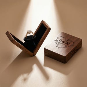 May include: A wooden ring box with a gold engagement ring inside, set against a black velvet interior. The box is open, displaying the ring. A closed box with a heart and floral design is next to it. Text includes names and a date.