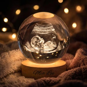 Peut inclure: Une boule de cristal transparente avec une image d'échographie d'un fœtus illuminée à l'intérieur. La boule repose sur un socle en bois gravé du nom "Mia Lee". La base est éclairée et l'arrière-plan est une texture douce et floue.