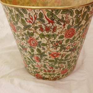 May include: A decorative waste bin with a floral and bird pattern. The design features red roses, green leaves, and birds on a cream background. The rim is trimmed with gold.