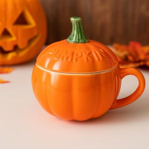 May include: Orange ceramic pumpkin-shaped mug with a green stem-shaped handle and lid. The word "PUMPKIN" is embossed on the lid. A carved pumpkin is in the background. Perfect for Halloween or fall.