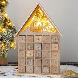 Puede incluir: Calendario de Adviento de madera con forma de casa con 24 cajones pequeños. El techo presenta un diseño recortado con una escena invernal retroiluminada de árboles, casas y un ciervo. Los cajones tienen pequeños pomos de madera.