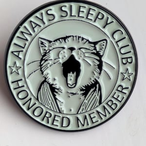 May include: A circular, mint-green enamel pin with a black border. The pin features a yawning cat illustration in the centre, surrounded by the words "ALWAYS SLEEPY CLUB HONORED MEMBER". Two stars flank the cat.