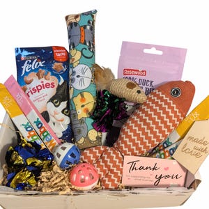 May include: A cat gift basket containing toys and treats. The basket includes a fish-shaped toy, a catnip tube, a mouse toy, and various treat packets. There are also two toy balls and a thank you card.
