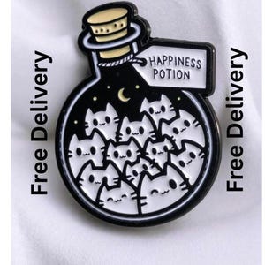 May include: A black and white enamel pin shaped like a round glass bottle filled with multiple small white cat faces. The bottle has a cork stopper and a white tag attached to the neck with the text HAPPINESS POTION printed in black letters.
