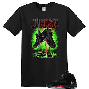 May include: A black short-sleeve t-shirt featuring a graphic of two black and red sneakers hanging by their laces. Above the sneakers is the word JORDAN in red text. Below the sneakers is a green splatter effect with red liquid dripping down.