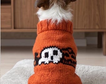 May include: A dog wearing a chunky knit orange sweater featuring a white and black skull graphic on the back. The sweater has a ribbed turtleneck collar and a ribbed hem, with the skull design centered horizontally across the mid-section.