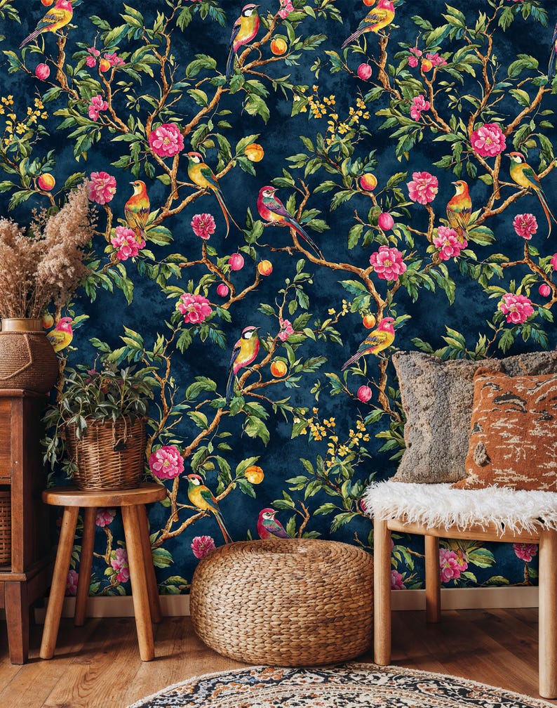 Cozy living room with navy blue chinoiserie wallpaper showing birds and floral branches. Natural wood accents, a rattan chair, pampas grass, and textured cushions enhance the bold botanical mural, blending vintage charm with modern bohemian style.