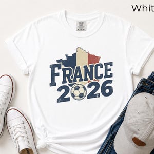 France 2026 soccer shirt with a vintage map of France in French flag colors. White Comfort Colors t-shirt mockup with retro distressed typography and a soccer ball graphic. Trendy world cup football fan gear flat lay with sneakers.