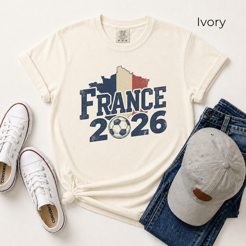 France 2026 soccer shirt with a vintage map of France in French flag colors. Ivory Comfort Colors t-shirt mockup with retro distressed typography and a soccer ball graphic. Trendy world cup football fan gear flat lay with sneakers.