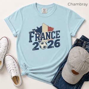 France 2026 soccer shirt with a vintage map of France in French flag colors. Chambray Comfort Colors t-shirt mockup with retro distressed typography and a soccer ball graphic. Trendy world cup football fan gear flat lay with sneakers.