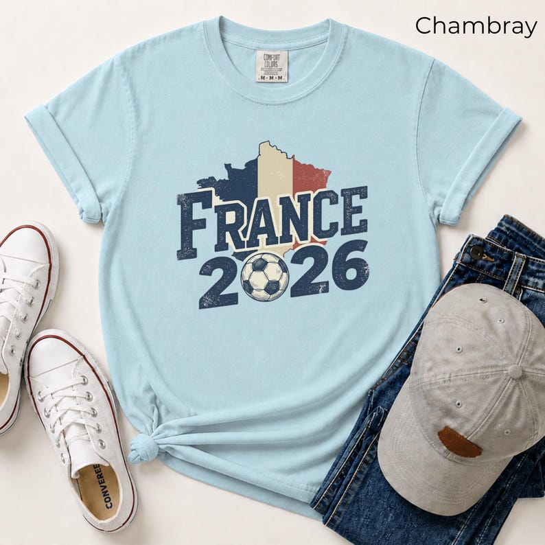 France 2026 soccer shirt with a vintage map of France in French flag colors. Chambray Comfort Colors t-shirt mockup with retro distressed typography and a soccer ball graphic. Trendy world cup football fan gear flat lay with sneakers.
